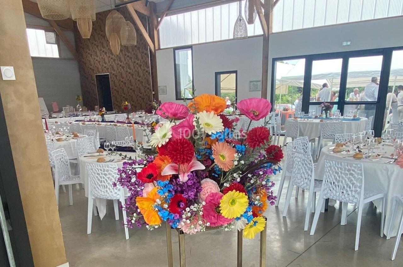 Composition florale colorée au premier plan dans une salle de réception lumineuse avec tables dressées en arrière-plan.