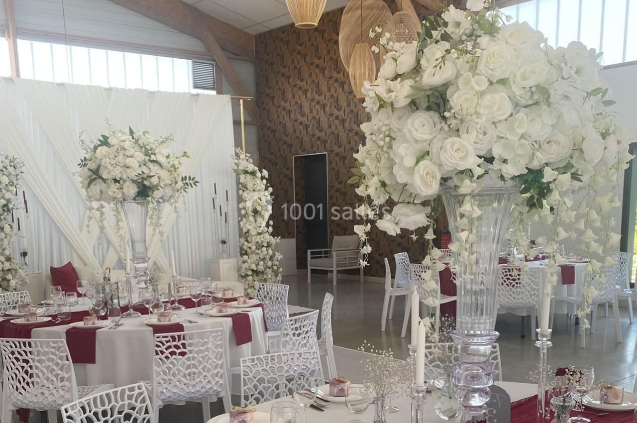 Salle de réception décorée avec des tables dressées, des chaises blanches ajourées et des compositions florales blanches.