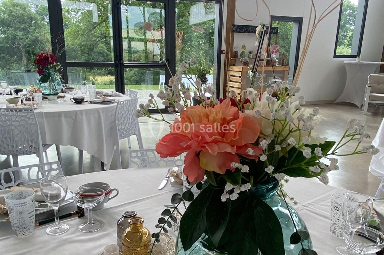 Salle de réception lumineuse avec tables décorées, centre de table floral et grandes baies vitrées donnant sur un jardin.