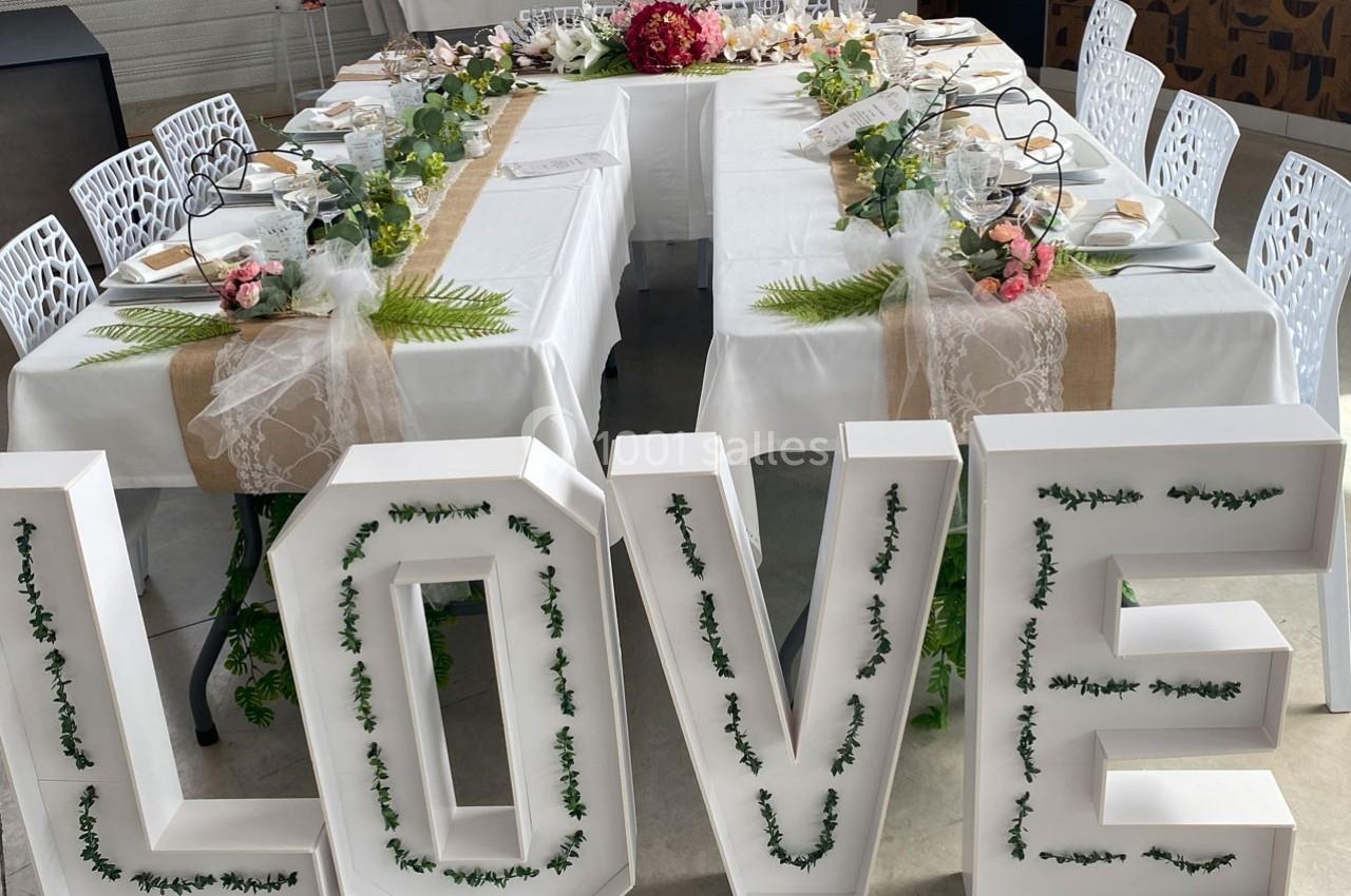 Table en U décorée pour un événement, ornée de fleurs, feuillages et lettres blanches formant le mot ’LOVE’.