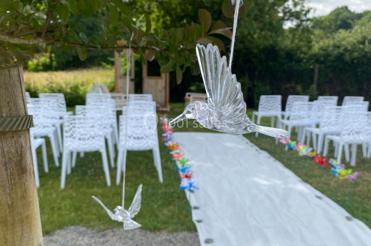 Décoration de mariage en extérieur avec des oiseaux en verre suspendus, allée blanche et chaises blanches alignées.