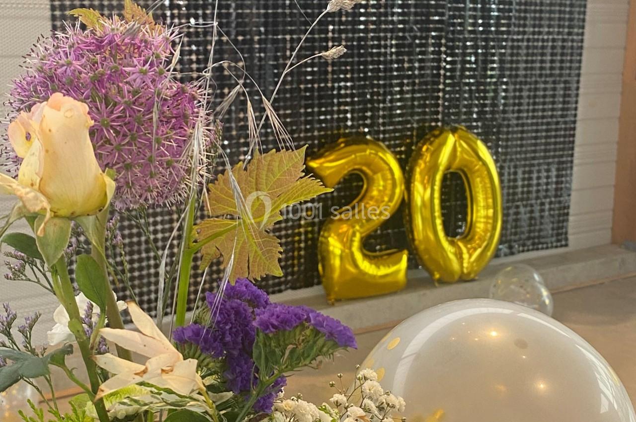 Ballons dorés formant le chiffre 20 devant un mur sombre, avec un bouquet de fleurs au premier plan.