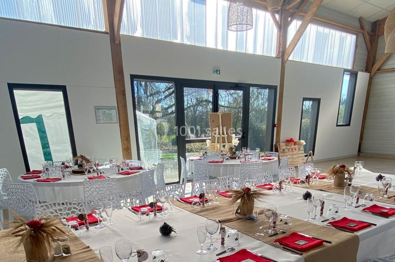 Salle de réception lumineuse avec tables dressées, nappes blanches et décorations champêtres rouges et beige.