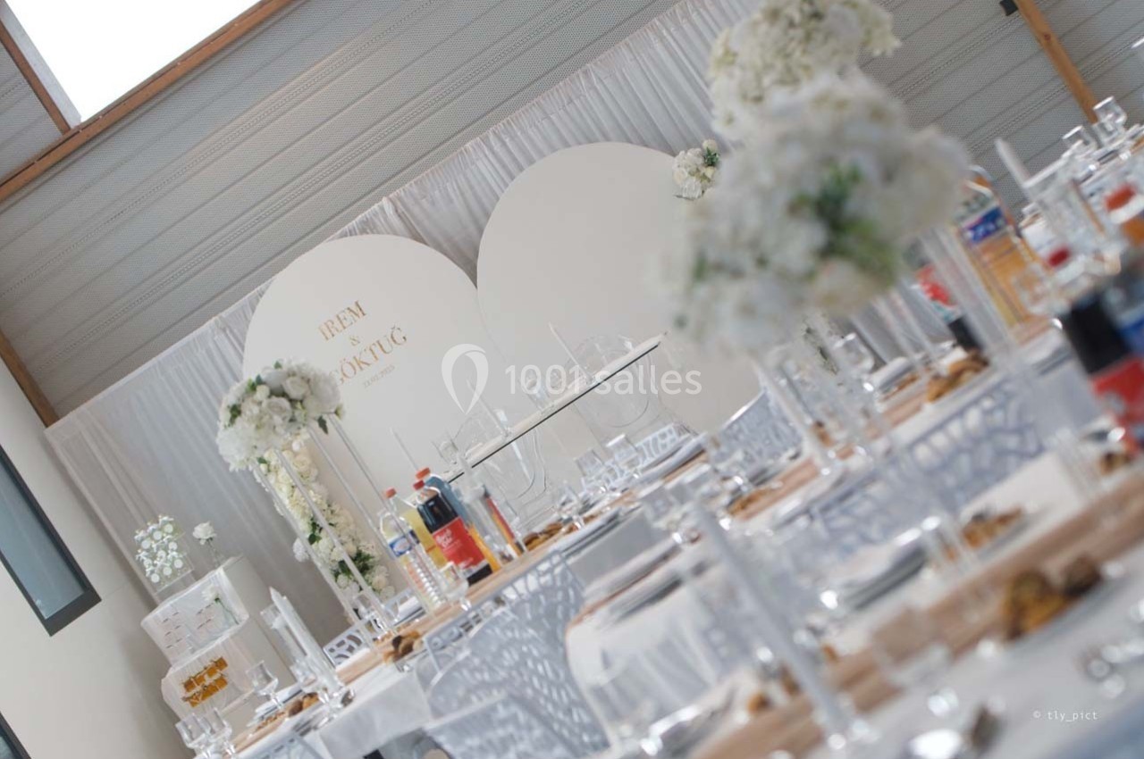 Salle décorée pour un événement avec des tables dressées, des chaises transparentes et des fleurs blanches en centre de…