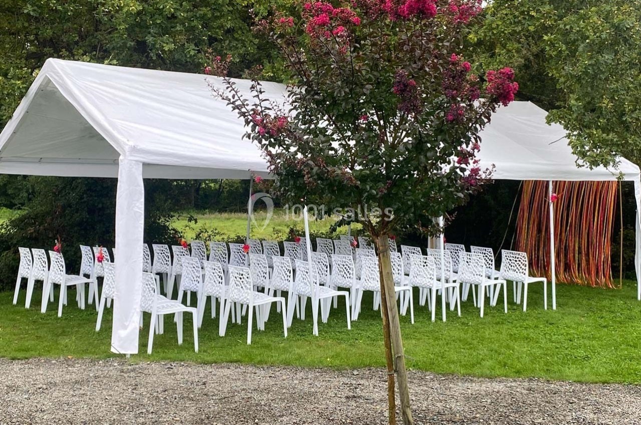 Tente blanche avec des chaises alignées pour un événement en extérieur, entourée de verdure et d'un arbre fleuri.
