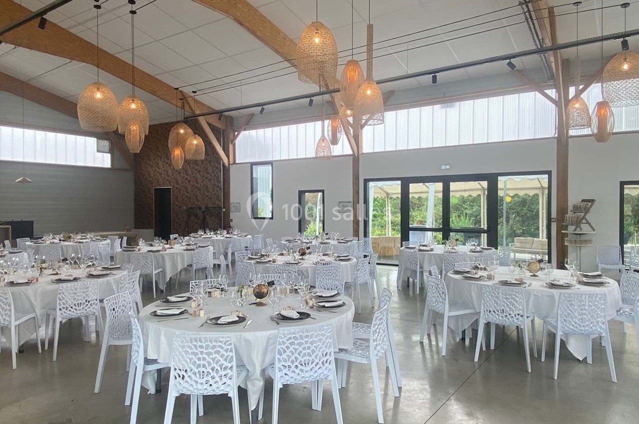 Salle lumineuse aménagée pour un événement, avec des tables rondes dressées et des chaises blanches ajourées.