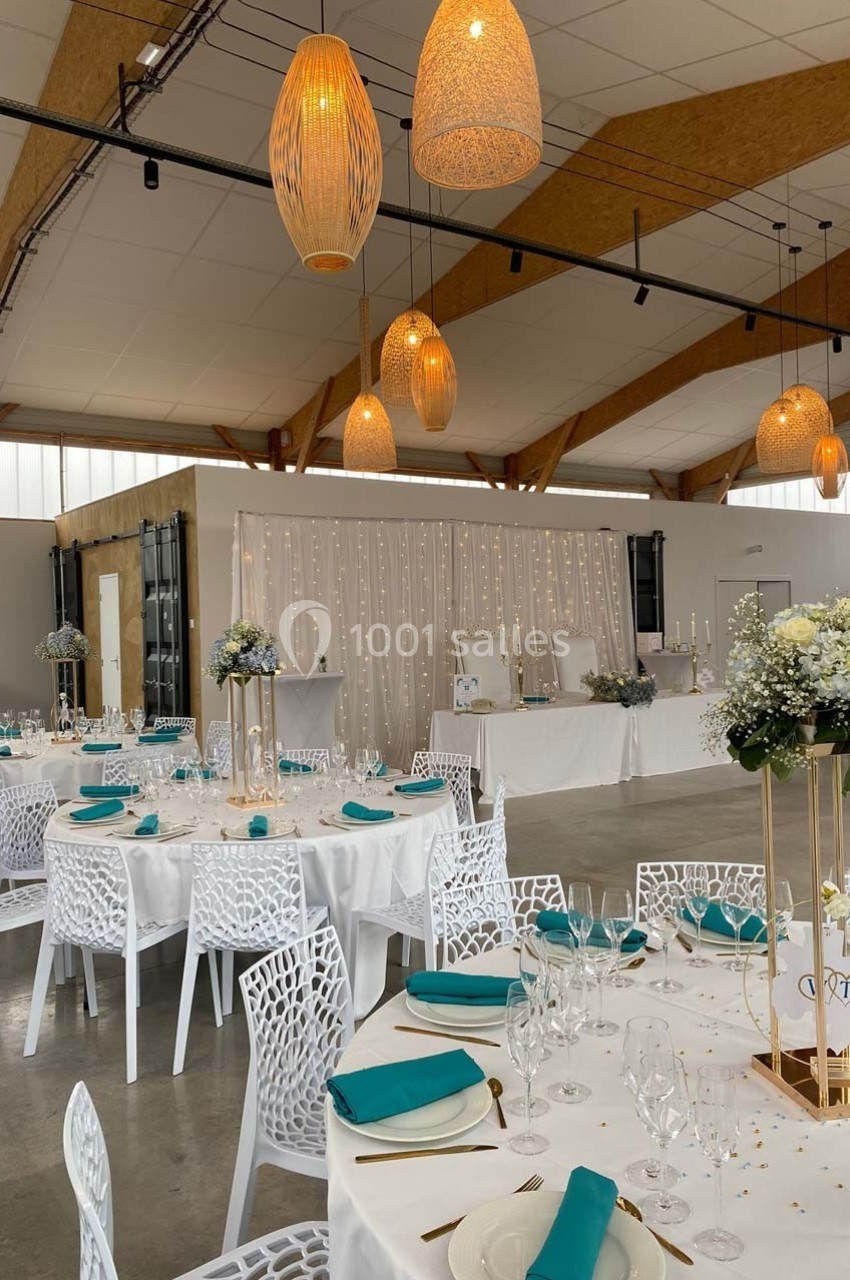 Salle de réception décorée avec des tables rondes, nappes blanches, serviettes turquoise et éclairage suspendu chaleureux.