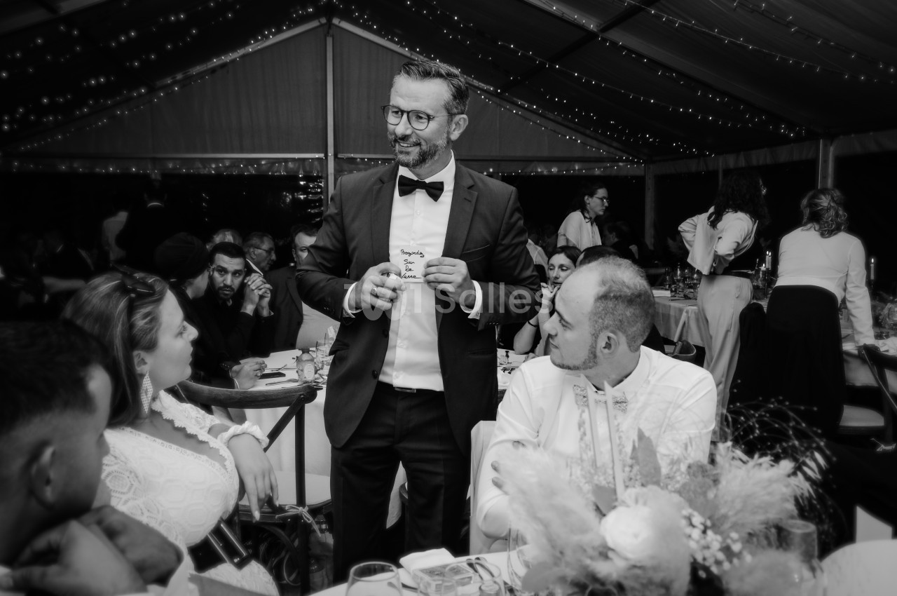 Un homme en costume parle à des invités assis autour d'une table lors d'un événement sous une tente éclairée.