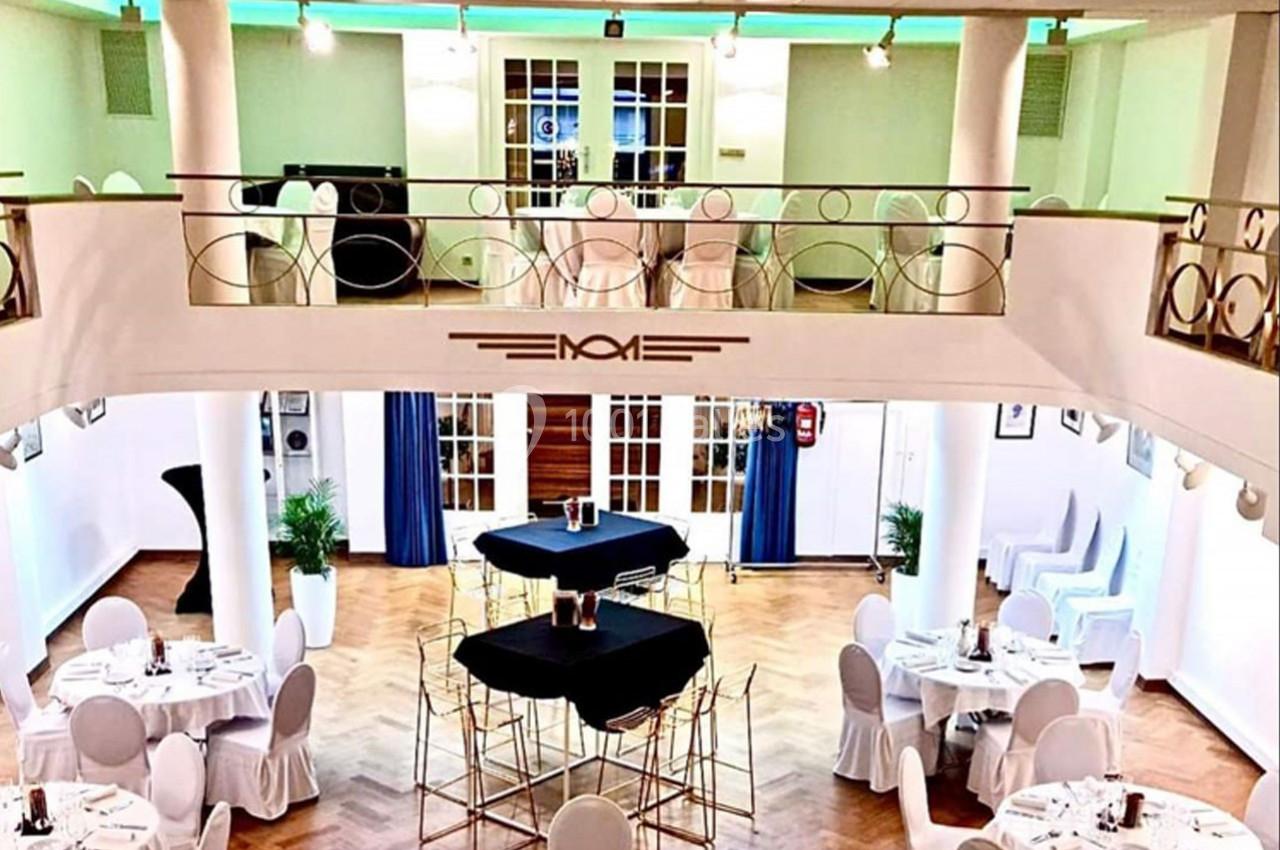 Salle de réception lumineuse avec mezzanine, tables rondes dressées et parquet en bois clair.