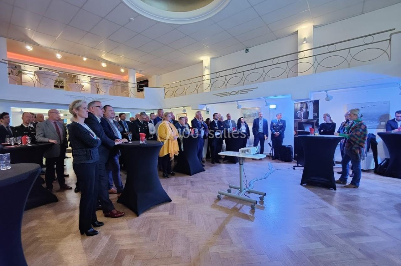 Groupe de personnes debout dans une salle lumineuse, participant à un événement avec des tables hautes et un projecteur.