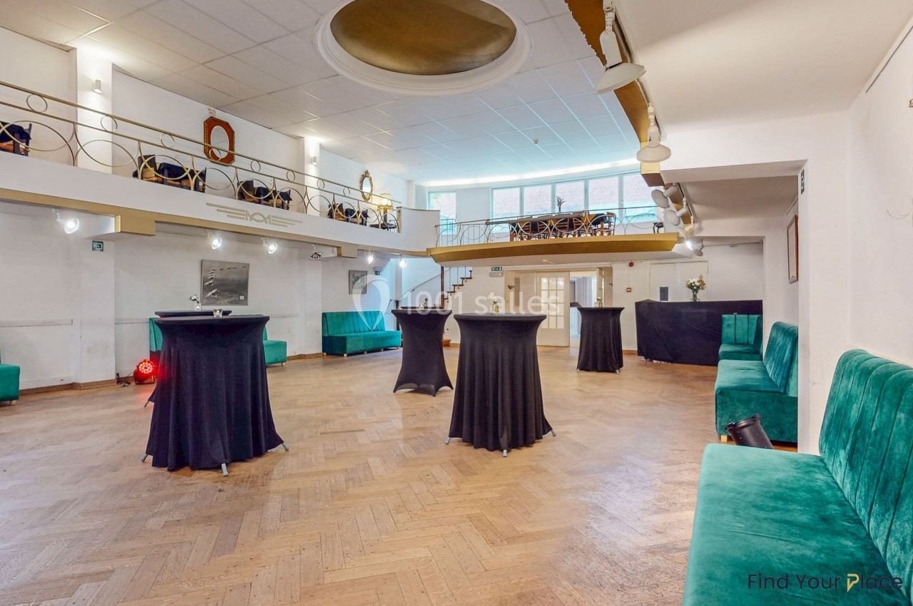 Salle lumineuse avec parquet en bois, tables hautes noires, banquettes vertes et mezzanine avec garde-corps doré.