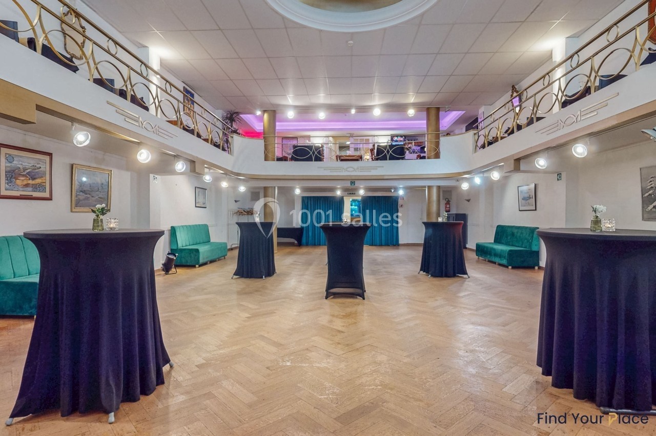 Salle lumineuse avec parquet, tables hautes drapées de noir, canapés verts et balustrade à l'étage.