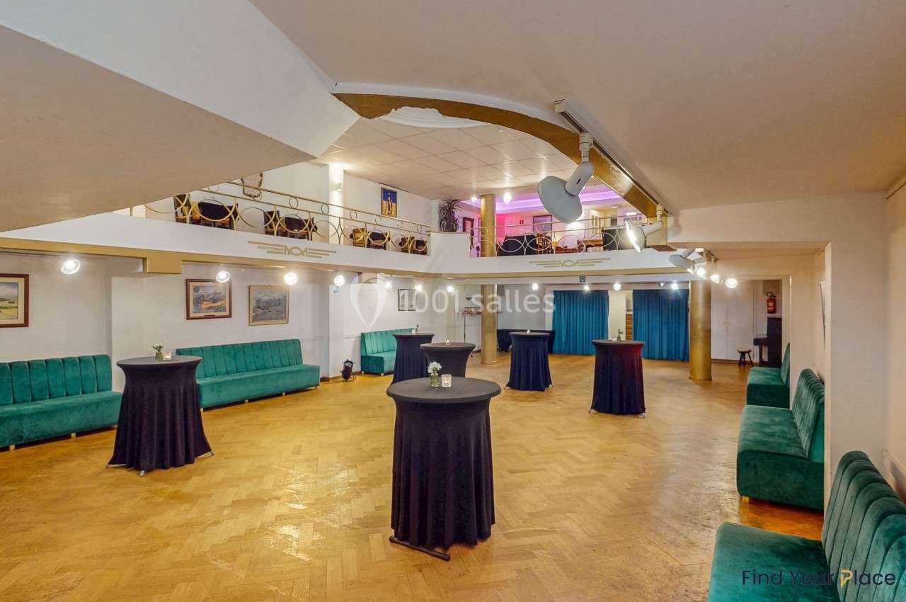 Salle lumineuse avec parquet, tables hautes noires, canapés verts et mezzanine avec sièges en arrière-plan.