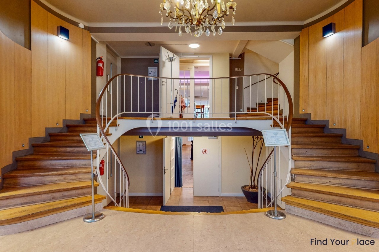 Escalier en bois à double volée dans un hall avec murs clairs, rampe métallique et lustre suspendu.
