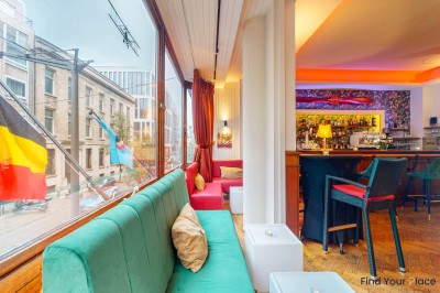 Intérieur d'un café lumineux avec canapés colorés, grandes fenêtres donnant sur une rue et un bar en arrière-plan.