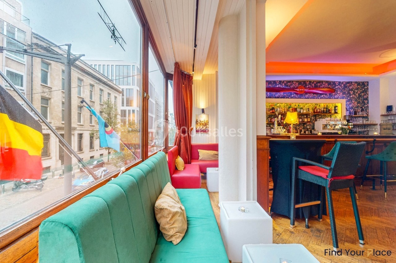 Intérieur d'un café lumineux avec canapés colorés, grandes fenêtres donnant sur une rue et un bar en arrière-plan.
