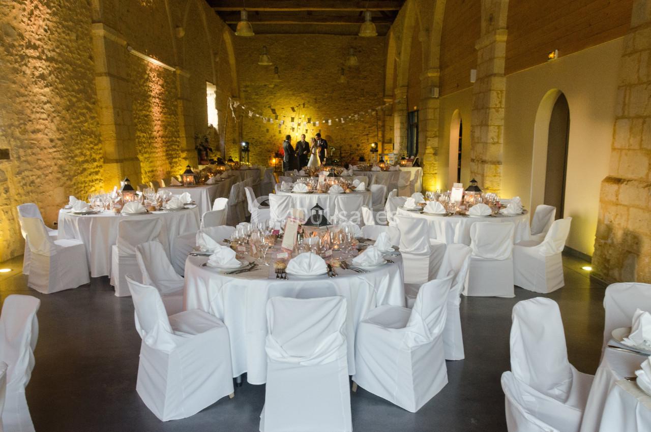 Salle de réception en pierre avec tables rondes dressées, nappes blanches et éclairage chaleureux.