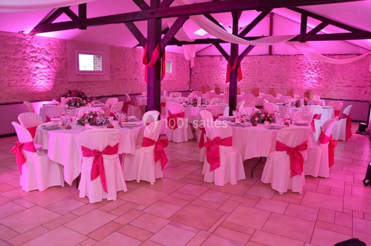 Salle de réception décorée avec des tables rondes, nappes blanches, chaises ornées de nœuds roses et éclairage rose.
