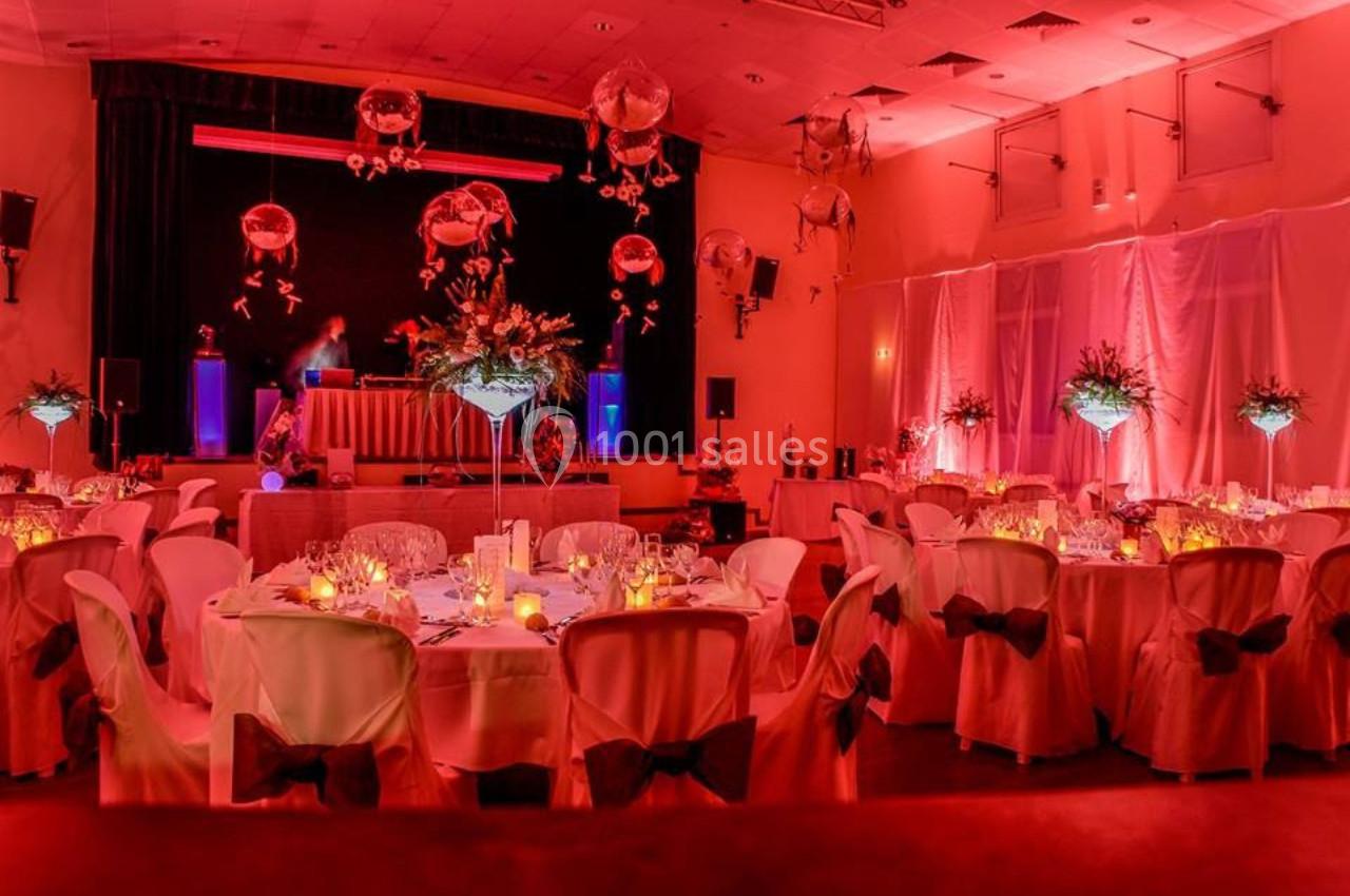 Salle de réception décorée avec des tables rondes, nappes blanches, éclairage rouge et arrangements floraux suspendus.