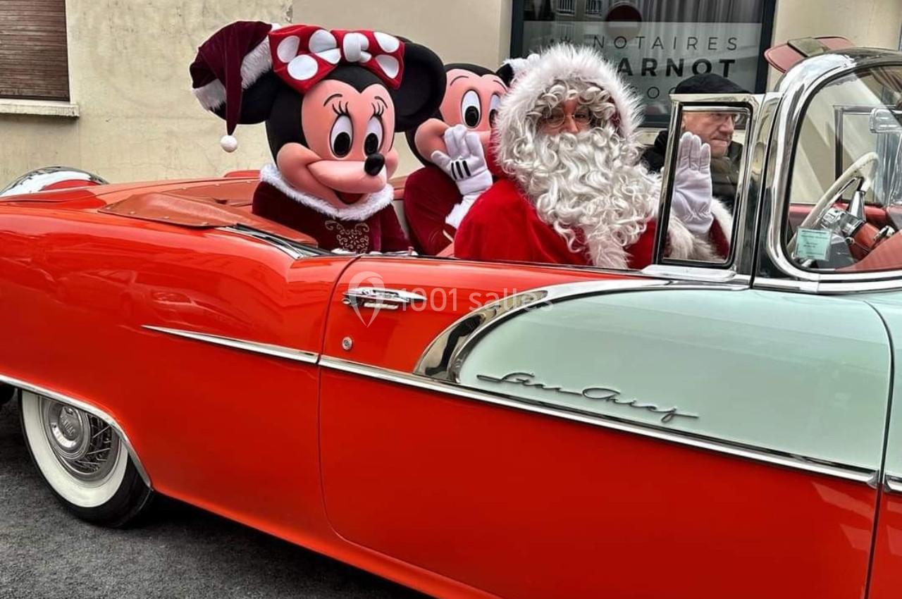 Le Père Noël et deux personnages costumés dans une voiture ancienne rouge et blanche, stationnée devant un bâtiment.