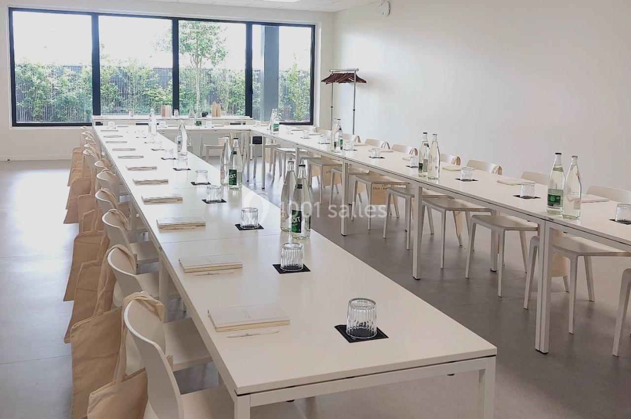 Salle lumineuse aménagée avec de longues tables blanches, des chaises, des bouteilles d'eau et des carnets disposés.