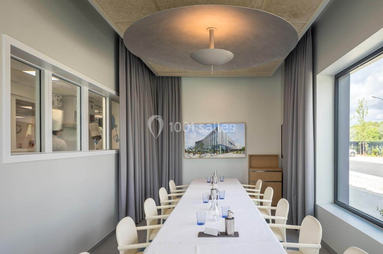 Salle de réunion lumineuse avec une grande table blanche, des chaises modernes, des rideaux gris et une vue extérieure.