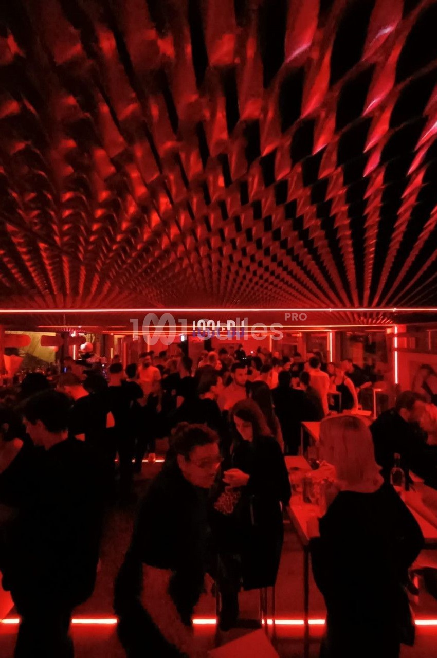 Salle de fête animée avec éclairage rouge, plafond texturé et personnes discutant ou dansant.