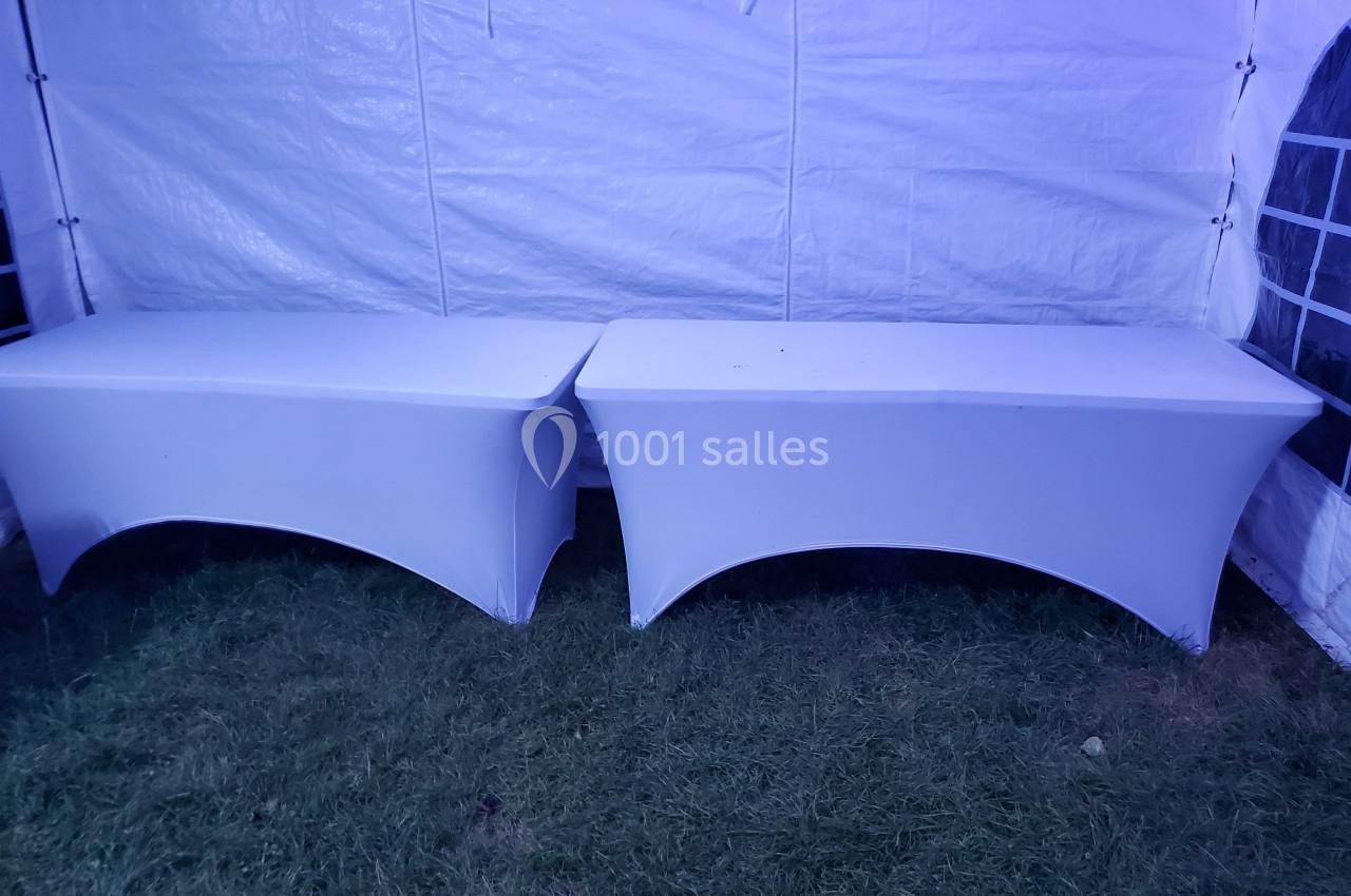 Deux tables rectangulaires recouvertes de housses blanches, placées côte à côte sur une pelouse sous une tente.