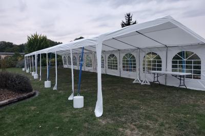 Grande tente blanche rectangulaire installée sur un parking, entourée de végétation et de barrières de sécurité.