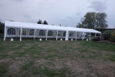 Grande tente blanche rectangulaire installée sur un parking, entourée de végétation et de barrières de sécurité.