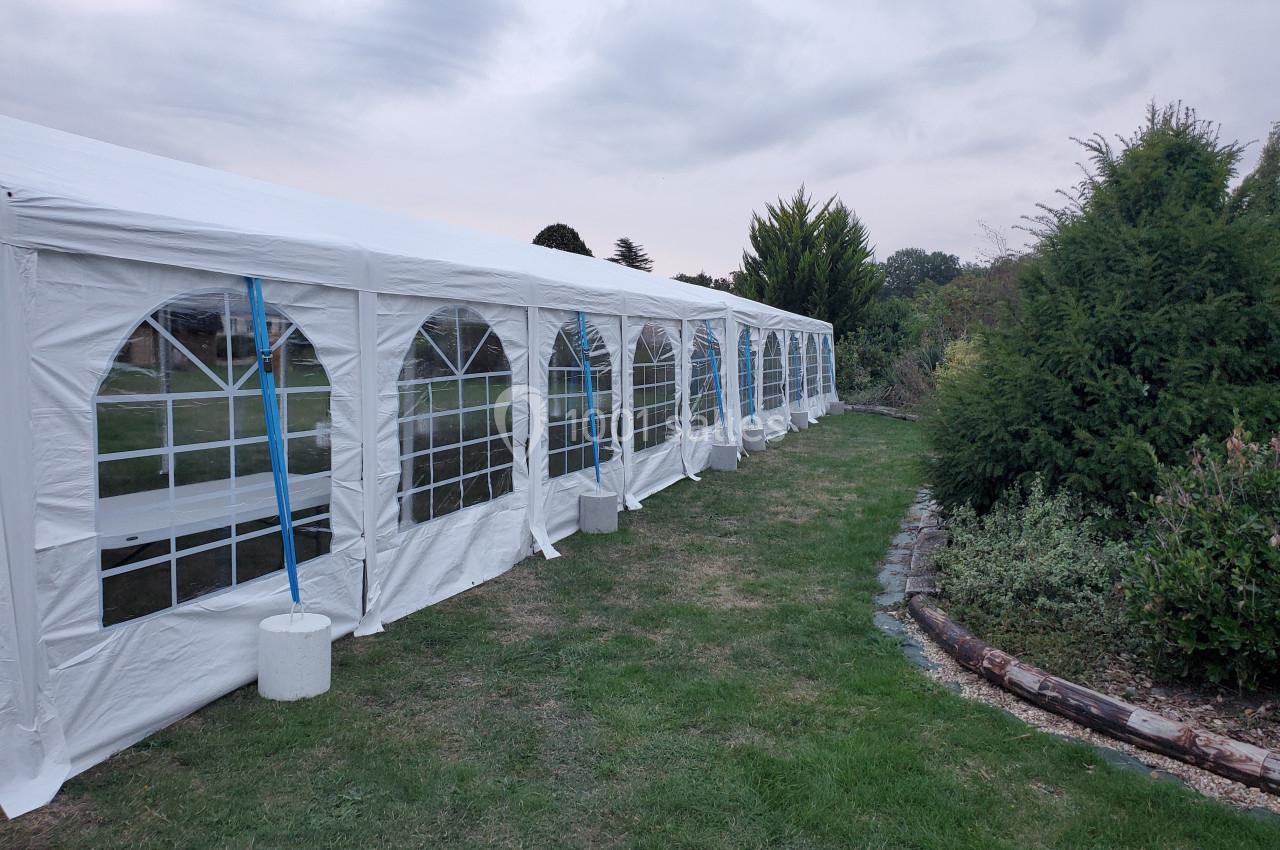 Grande tente blanche avec fenêtres en arc, installée sur une pelouse près d'arbustes et d'un ciel nuageux.