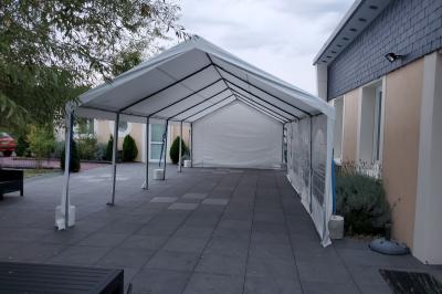 Grande tente blanche rectangulaire installée sur un parking, entourée de végétation et de barrières de sécurité.