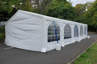 Grande tente blanche rectangulaire installée sur un parking, entourée de végétation et de barrières de sécurité.