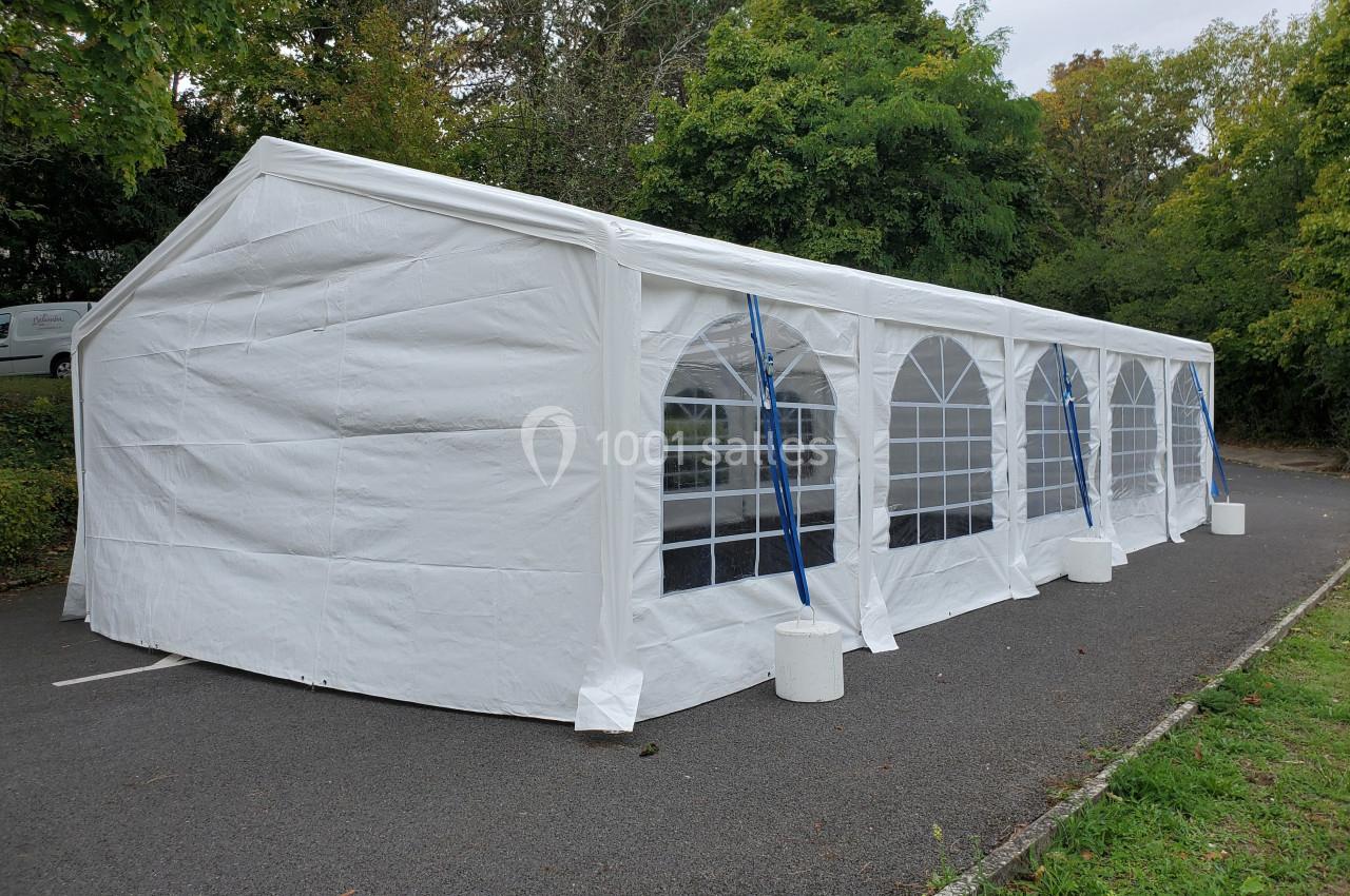 Grande tente blanche avec fenêtres en arc, installée sur un parking entouré de végétation.