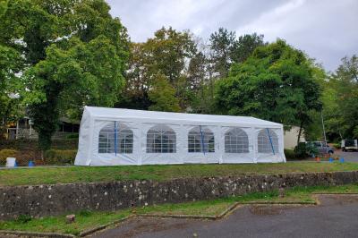 Grande tente blanche rectangulaire installée sur un parking, entourée de végétation et de barrières de sécurité.
