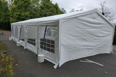 Grande tente blanche rectangulaire installée sur un parking, entourée de végétation et de barrières de sécurité.