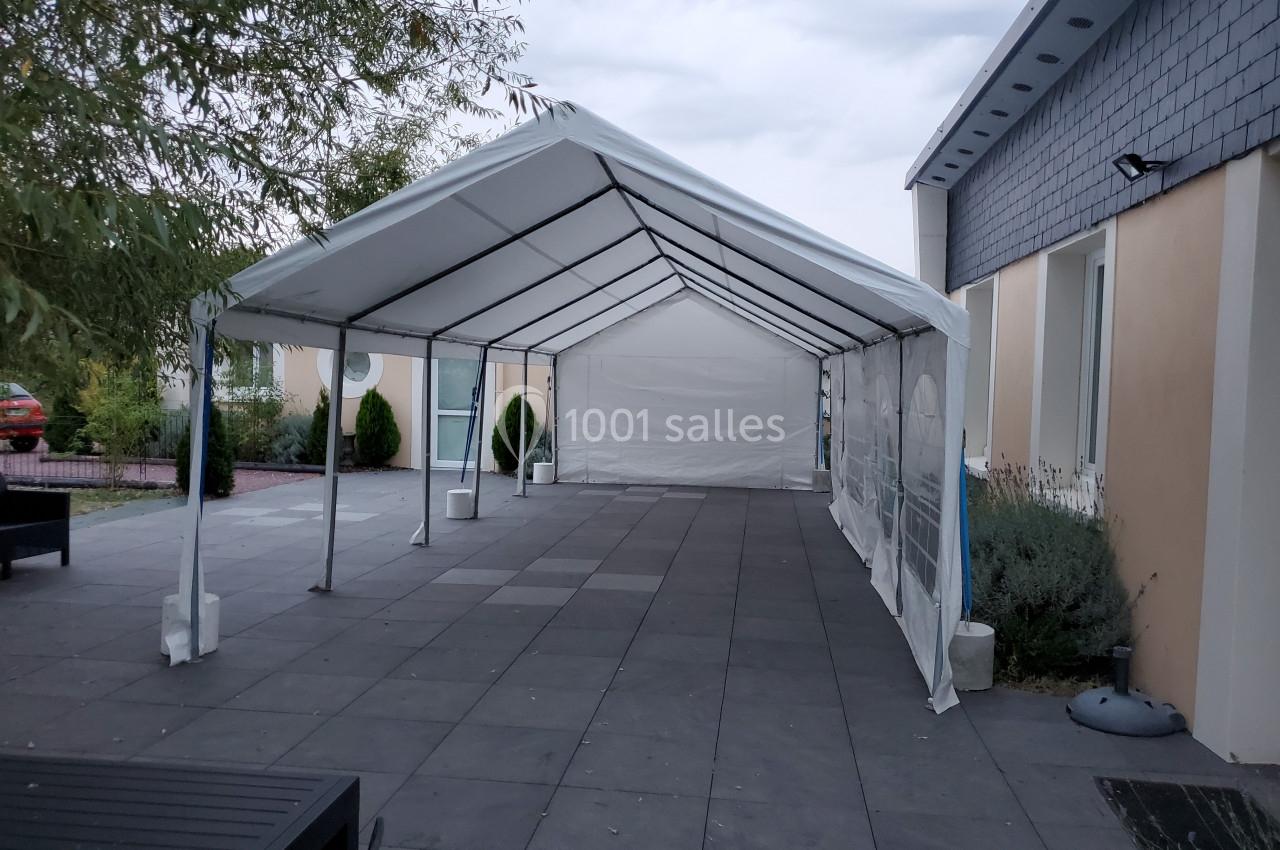 Tente blanche montée sur une terrasse pavée, entourée de bâtiments et d'arbres sous un ciel nuageux.