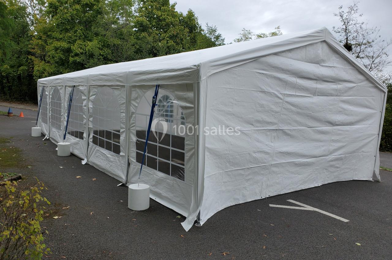 Grande tente blanche rectangulaire installée sur un parking, avec des parois transparentes et des arbres en arrière-plan.