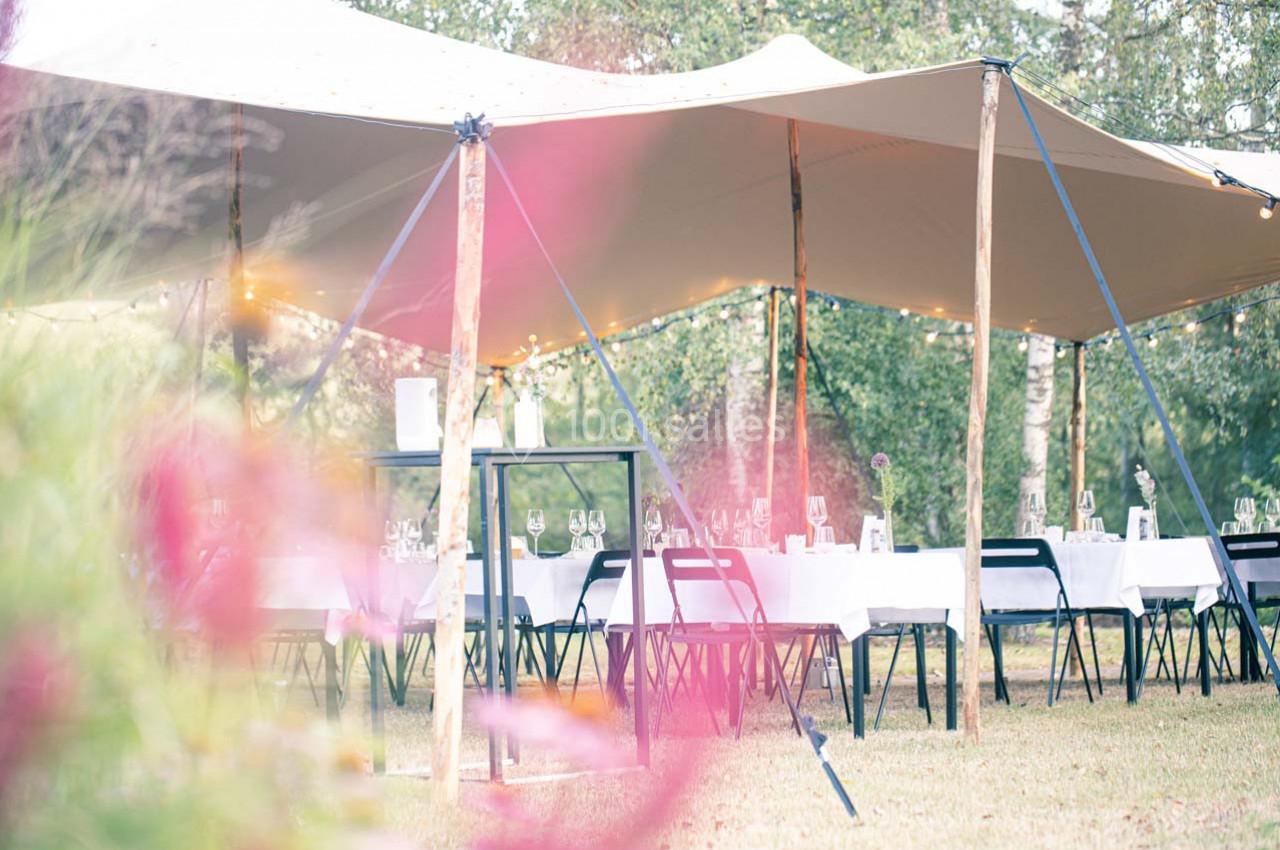 Tables dressées sous une tente beige dans un jardin, avec des fleurs floues au premier plan.