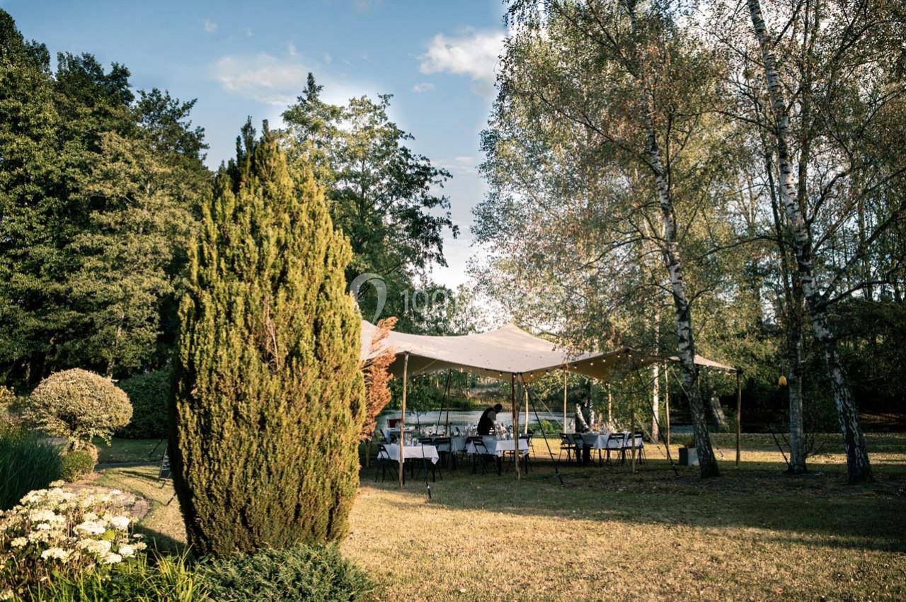 Tente beige installée dans un jardin arboré, avec des tables dressées pour un événement en plein air.