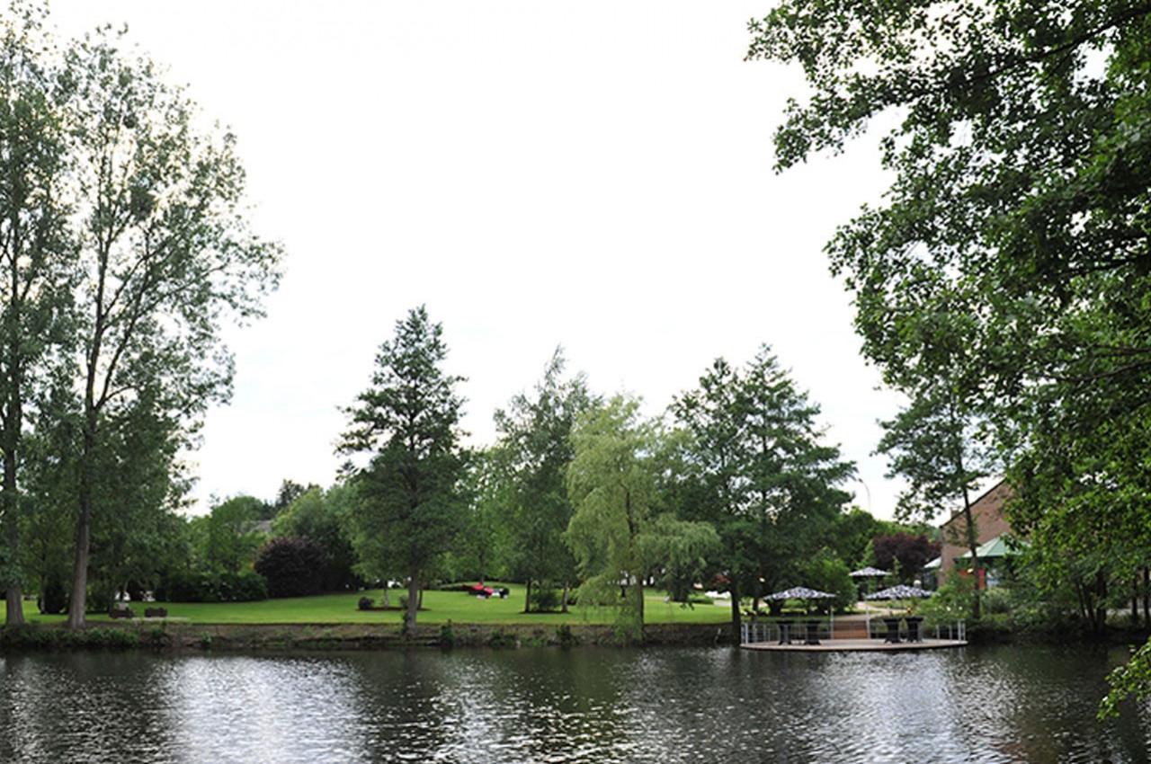 Vue d'un parc verdoyant avec des arbres, une pelouse et une rivière au premier plan, sous un ciel clair.