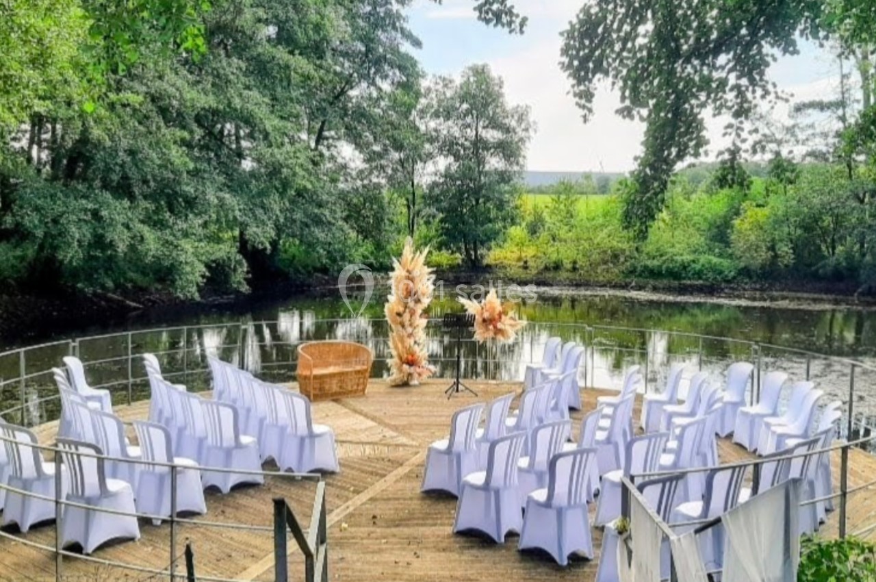 Chaises blanches disposées en cercle sur une plateforme en bois au bord d'un étang, entourée de végétation.
