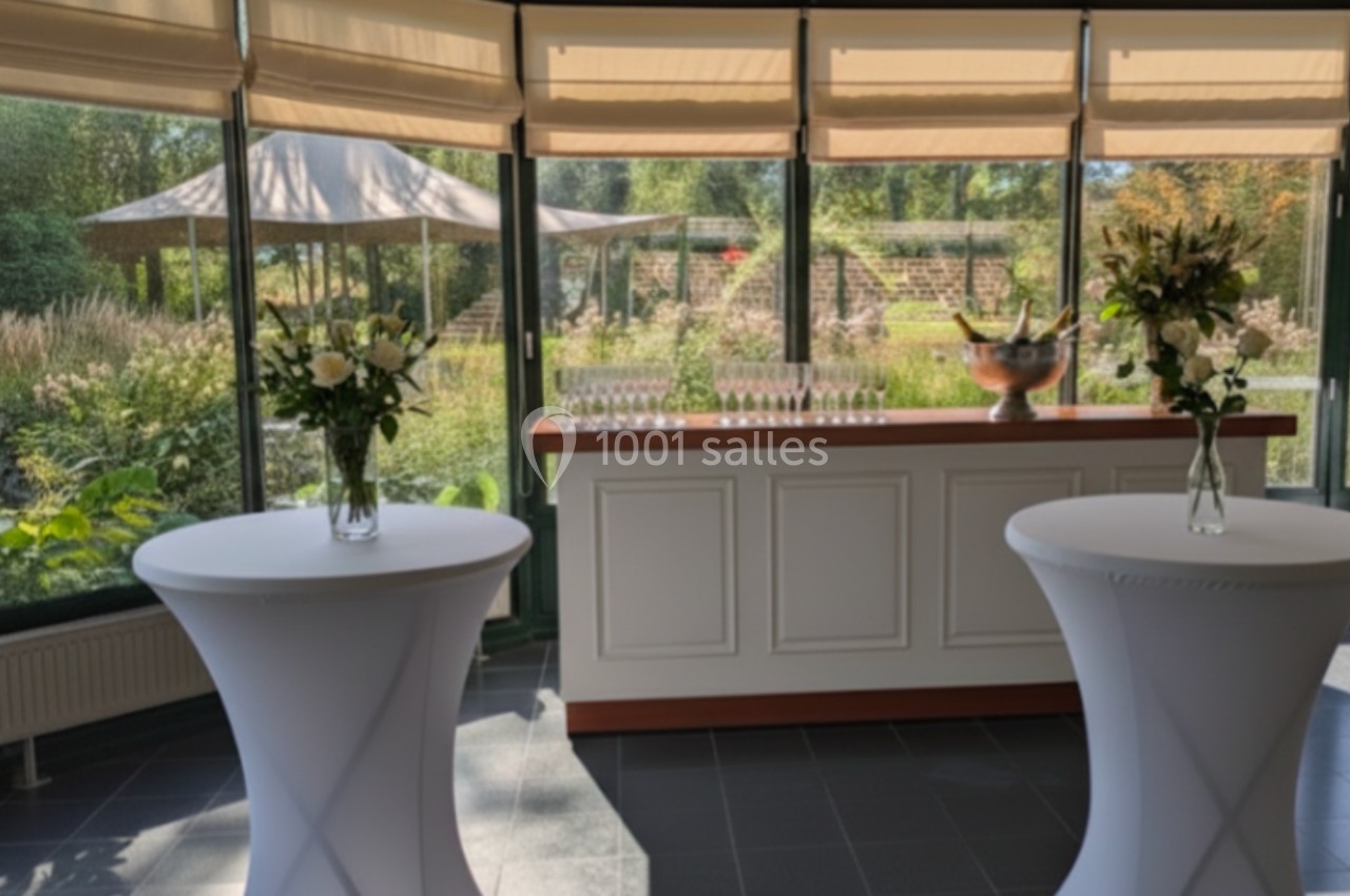 Salle lumineuse avec baies vitrées, deux tables hautes décorées de fleurs et un bar en arrière-plan.