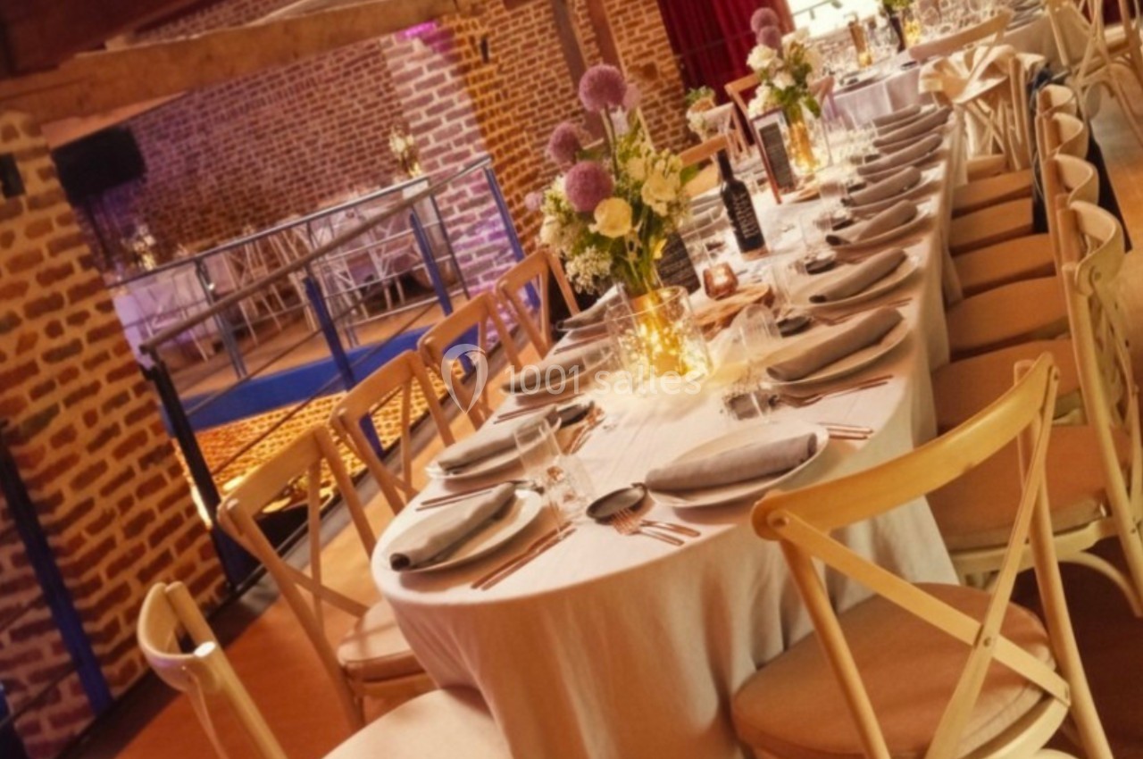 Salle de réception avec tables rondes dressées, nappes blanches, chaises en bois et décorations florales.