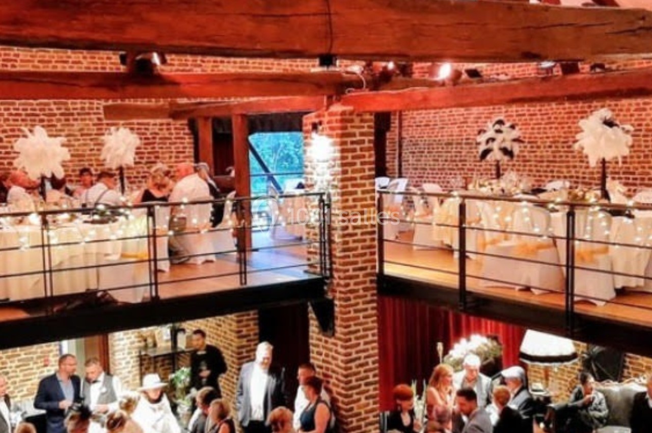Salle de réception en briques avec mezzanine, décorée pour un événement, invités debout et tables dressées.