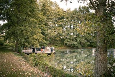 Jardin verdoyant avec arbres, pelouse soignée, tables hautes sous parasols et mobilier coloré près d'un plan d'eau.