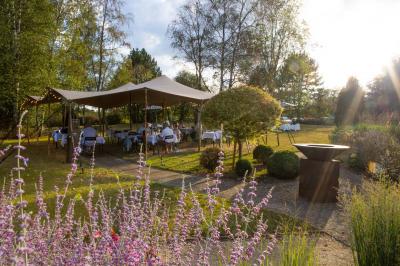 Jardin verdoyant avec arbres, pelouse soignée, tables hautes sous parasols et mobilier coloré près d'un plan d'eau.