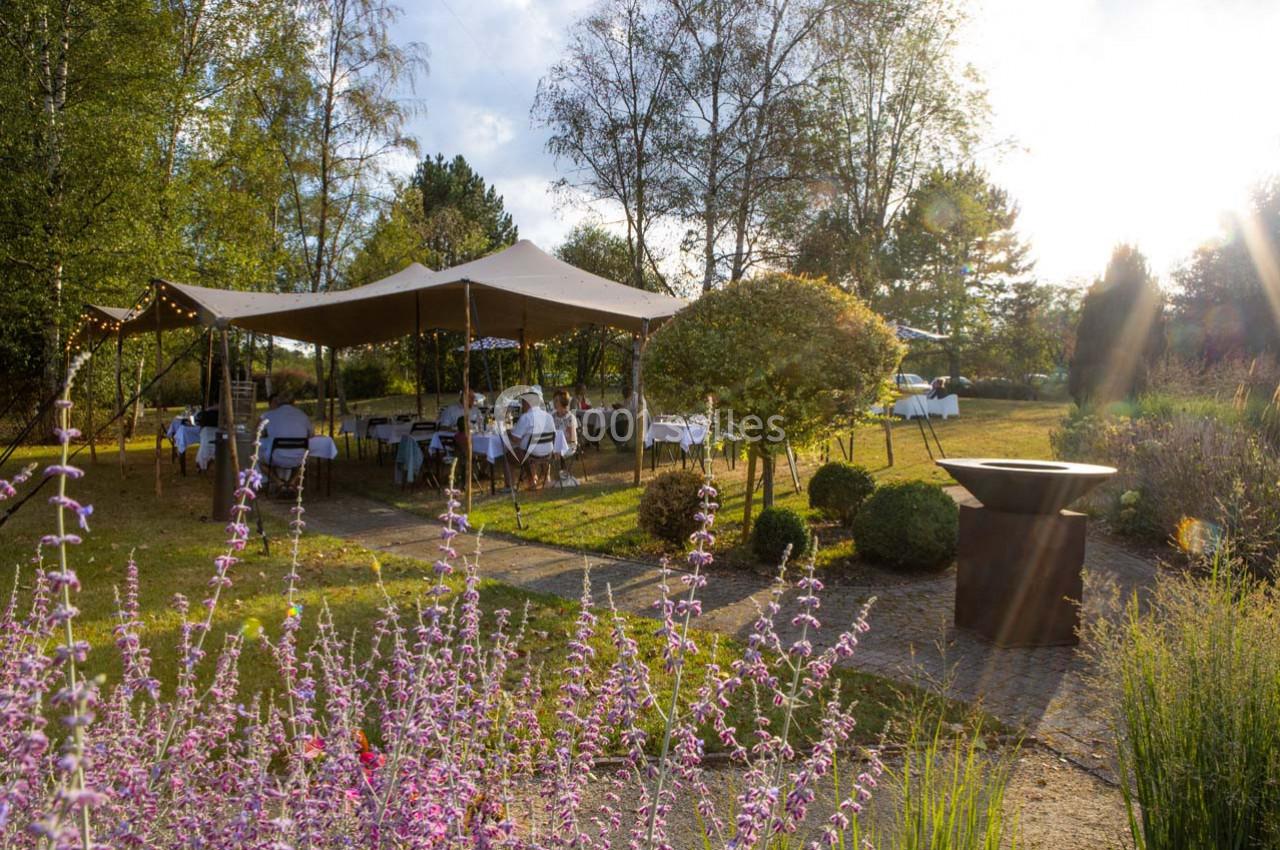 Tente installée dans un jardin verdoyant avec des tables dressées, entourée de fleurs et d'arbres sous un soleil couchant.