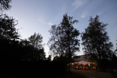 Jardin verdoyant avec arbres, pelouse soignée, tables hautes sous parasols et mobilier coloré près d'un plan d'eau.