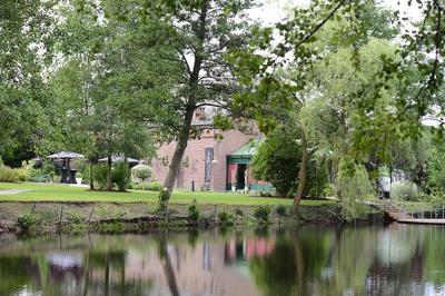 Jardin verdoyant avec arbres, pelouse soignée, tables hautes sous parasols et mobilier coloré près d'un plan d'eau.